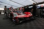 Red and black race car with open doors in a garage surrounded by crew members and equipment.