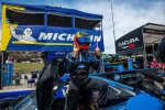 Race car driver in blue suit celebrating atop Acura car with Michelin and Acura banners in background.