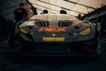 Front view of a matte black Lamborghini race car with yellow headlights and DEX logo on hood in racing pit.