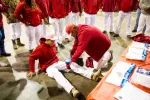 Group in red hoodies and white pants, one person helps seated man with leg injury indoors with medical supplies nearby