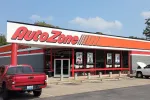 AutoZone store exterior with red signage, parked trucks, and promotional posters on windows under a sunny sky