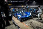 Blue Acura race car in night pit stop with crew changing tires and refueling on illuminated racetrack.