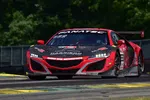Red and black racing car with Fanatec logo speeding on racetrack surrounded by green trees under bright daylight