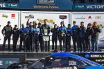 Racing team celebrates winning at Daytona International Speedway Victory Lane with trophy and blue race car.