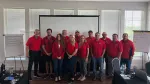 Team of twelve people wearing red shirts standing in a conference room with presentation boards and a projector screen.