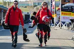 Two race car drivers in red jackets walking with helmets and gear at a racing event pit lane.