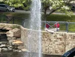 Water fountain sprays high above a stone retaining wall with people and greenery in the background