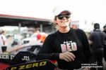 Smiling female race car driver in sunglasses and visor stands beside P Zero branded race car at racetrack.