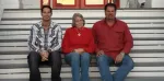 Three adults sitting on wooden steps outdoors, two men and one woman in casual attire smiling at camera.