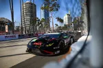 Black Lamborghini race car on city street circuit with high-rise buildings and safety fencing in background