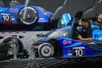 Mechanic working on blue racing car number 10 in garage with car parts and trailer in background