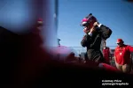 Race car driver adjusts helmet preparing for event with crew members and a clear blue sky in background.