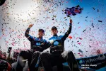 Two joyful race car drivers in blue uniforms celebrate victory with confetti and a waving checkered flag under bright sunlight.