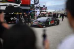 Black and red race car number 15 at a racetrack pit lane with crew members and spectators nearby.
