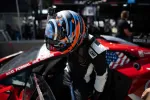 Race car driver in orange and blue helmet entering a red racing car with American flag decal on door.