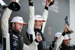 Three race car drivers celebrating on the podium holding trophies and wearing racing suits and caps.