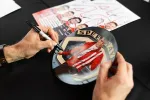 Person signing a collectible plate featuring a man in a red and white striped jacket with a black marker.