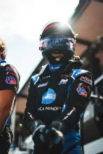 Race car driver in black and blue racing suit and helmet preparing in pit area with team member beside them.