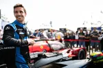 Smiling race car driver in blue and black suit standing next to a red and black race car with crowd in background