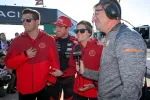 Race car driver in black suit with two people in red jackets and a reporter conducting interview outdoors.