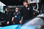 Three racing team members in black and blue uniforms standing in a garage with a race car and equipment around them.