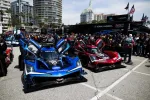 Two high-performance Acura race cars, one blue and one red, with open doors surrounded by crew and spectators.