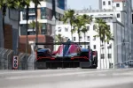 Rear view of a race car speeding on an urban street track with buildings and palm trees in the background
