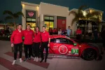 Four people in red shirts pose beside a red race car at night outside a lit modern building with palm trees.