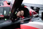 Race car driver waving while seated inside a red and black Acura DPi racing car at the track.