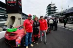 Racing driver and two team members posing next to a red Acura race car at a motorsport event pit lane.