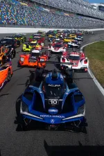 Wide shot of colorful prototype race cars lined up on a race track at Daytona with grandstands in the background
