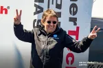Man in black jacket and sunglasses making peace signs with both hands at an outdoor event.