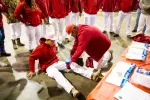 Person in red hoodie receives first aid on leg from another individual in red jacket and cap indoors.