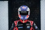 Race car driver in black Acura suit and colorful helmet standing between illuminated panels for dramatic effect.