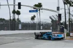 Blue and black race car number 10 speeding on a city street race track with Pine Ave sign and palm trees.