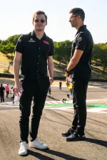 Two men in black Acura DEX racing team shirts standing on a racetrack on a sunny day holding drinks.