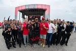 Race car drivers and team members celebrate a victory with raised index fingers at a motorsport event podium.