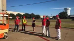Group of six workers wearing red helmets and safety gear standing outside near construction equipment under clear blue sky.