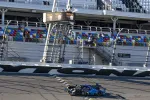 Race car crosses finish line at Daytona International Speedway with colorful empty grandstands in background