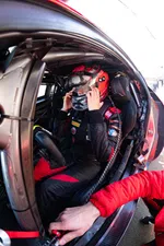 Race car driver wearing a red and black helmet and suit prepares inside the car with a crew member assisting.