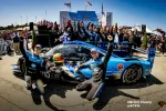 Race car drivers and team celebrate victory with blue Konica Minolta Acura race car at a motorsports event podium.