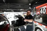 Bearded man wearing sunglasses working on car parts under a black canopy near a race truck marked 40