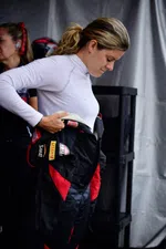 Race car driver wearing a white undershirt puts on a black and red racing suit in a garage area.