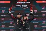 Two race car drivers in black suits and VP hats holding trophies on a podium at GT World Challenge America powered by AWS.