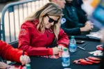 Woman in red jacket and sunglasses signing autographs at a table with bottled water and fans nearby