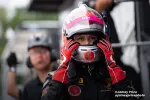 Female race car driver adjusting helmet with red gloves in the pit lane, focused and ready for the race.