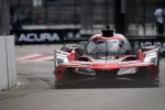 Red Acura prototype race car with bright headlights racing on a street circuit near a concrete barrier.