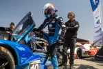 Race car driver in blue suit entering a Konica Minolta Acura prototype race car during a pit stop.