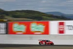 Red race car speeding on racetrack with blurred sponsor banners and green hills in the background
