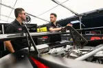 Two mechanics in black shirts discussing beside a race car's exposed rear wing and engine in a garage.
